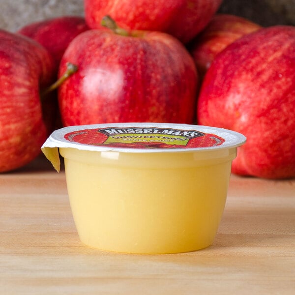 A yellow container of Musselman's unsweetened applesauce on a table with apples.