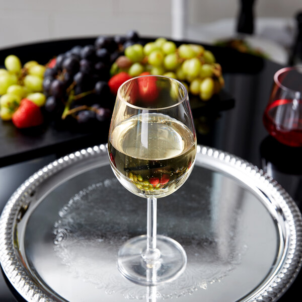 A customizable Tritan plastic tall wine glass filled with white wine on a tray with grapes and fruit.