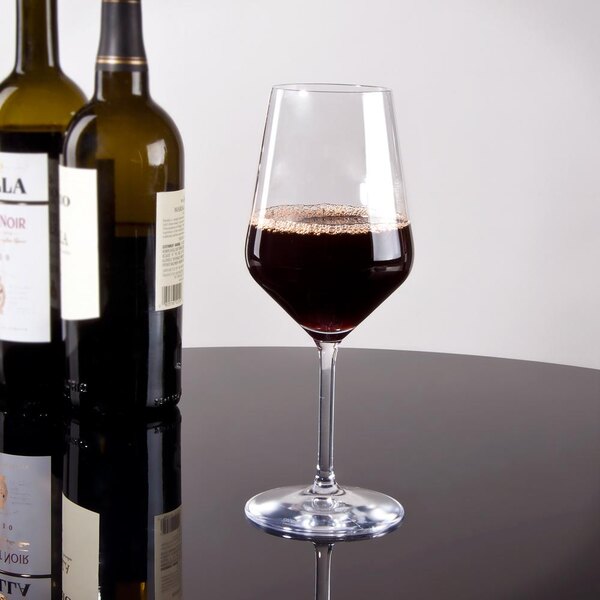 A Stolzle all-purpose wine glass filled with red wine on a table next to two bottles of wine.