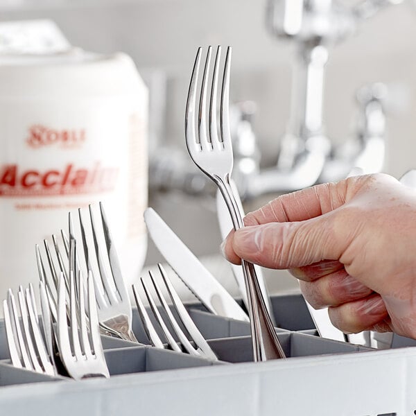 A hand holding a fork in front of a bottle of Noble Chemical Acclaim Concentrated Solid Dish Machine Detergent.