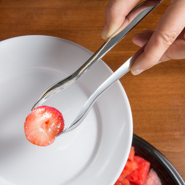 A person using Visions silver plastic tongs to pick up a piece of fruit.