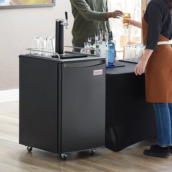 A woman and a man standing next to a black Galaxy Single Tap Kegerator Beer Dispenser on wheels.