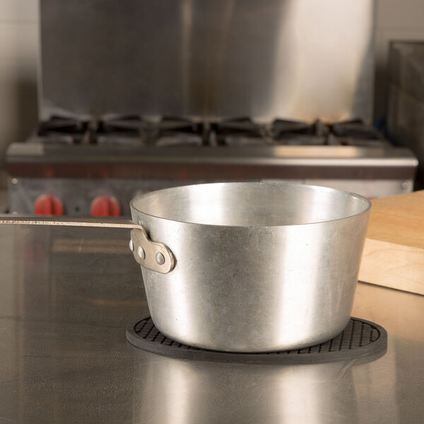 An American Metalcraft black silicone oval trivet holding a metal pot on a table.