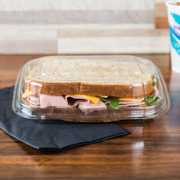 A sandwich in a 13 oz clear plastic take out container on a table in a deli.