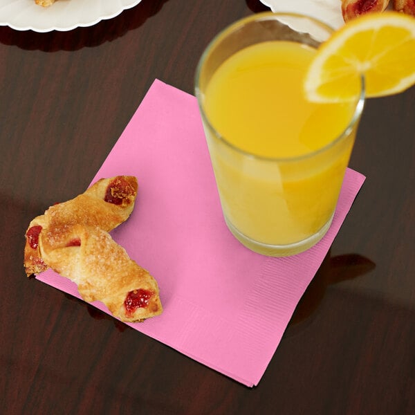 A glass of orange juice next to pastries on a pink Creative Converting beverage napkin.
