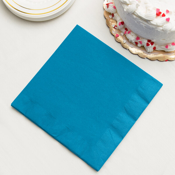 A turquoise blue Creative Converting paper dinner napkin on a stack of plates with a white cake on top.