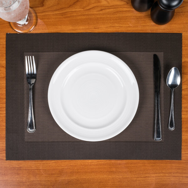 A white plate with a fork and spoon on a Snap Drape Reno chocolate PVC placemat on a brown table.