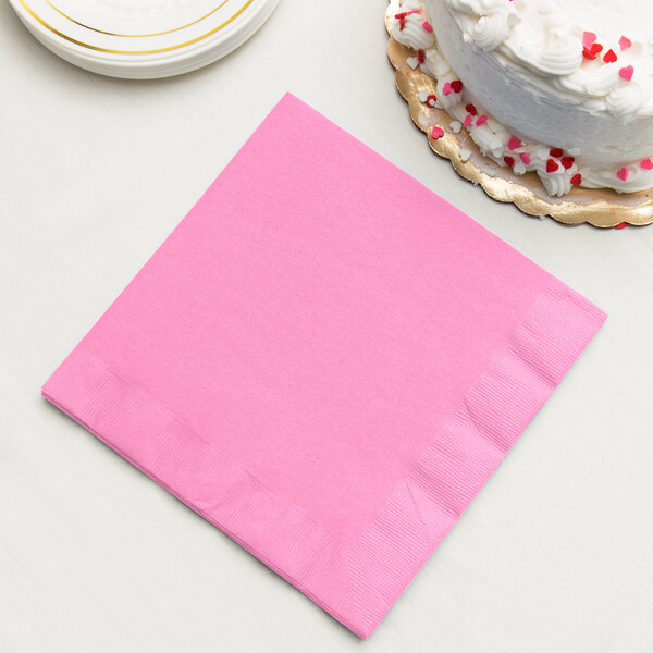 A pink Creative Converting 3-ply paper dinner napkin next to a white cake.