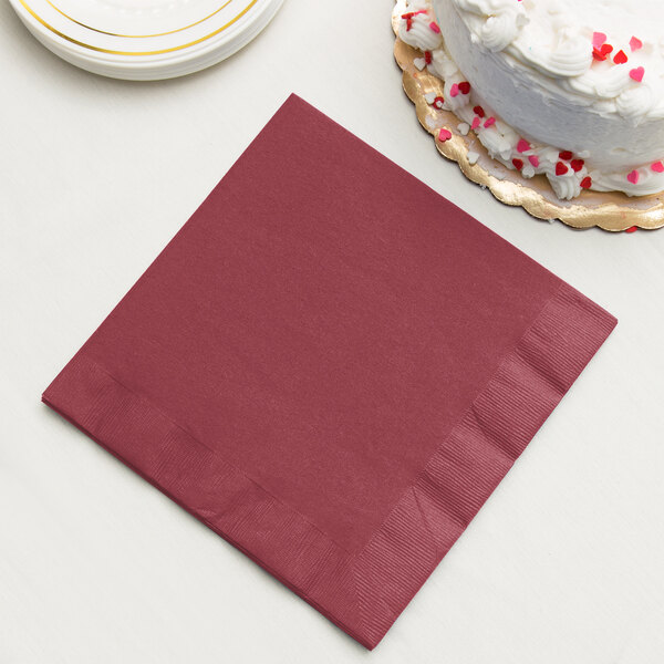 A burgundy Creative Converting 3-ply paper dinner napkin next to a cake.