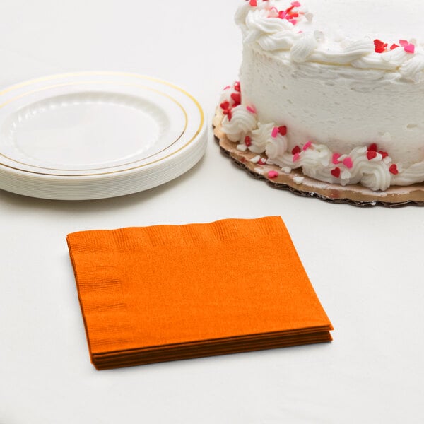 A stack of orange Creative Converting beverage napkins next to a white cake with red sprinkles.