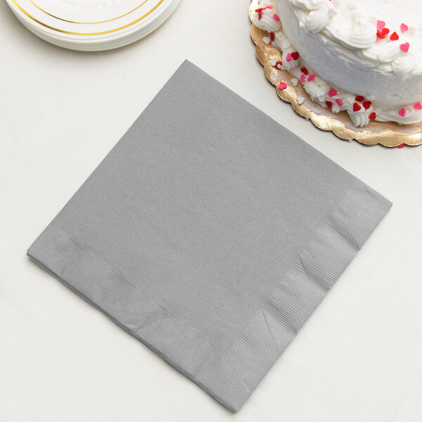 A Creative Converting shimmering silver paper dinner napkin on a table next to a white cake with red and pink sprinkles.