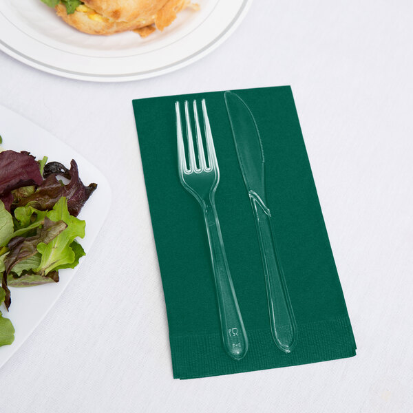 A fork and knife on a green napkin next to a plate of salad.