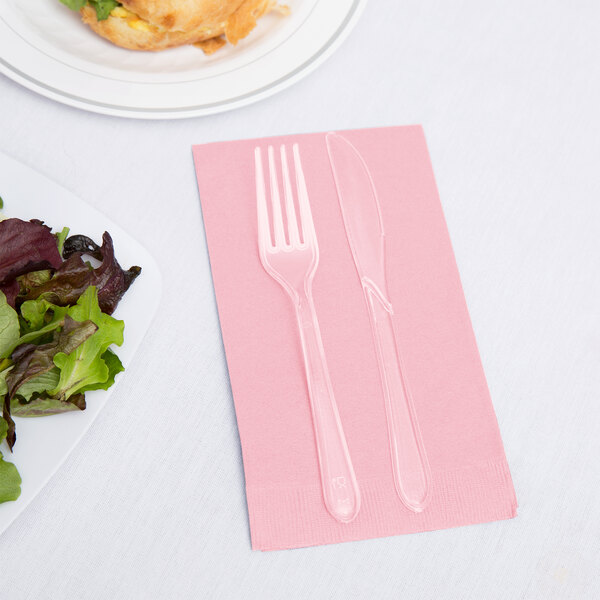A fork and knife on a pink napkin next to a plate of salad.