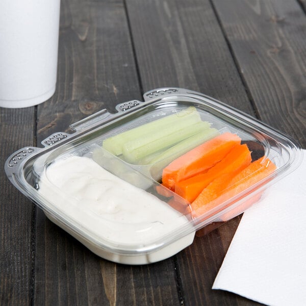 A 13 oz. clear plastic deli container with carrots, celery and cucumbers on a white table.