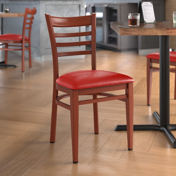 A Lancaster Table & Seating metal ladder back chair with mahogany wood grain and red vinyl seat on a table in a restaurant dining area.