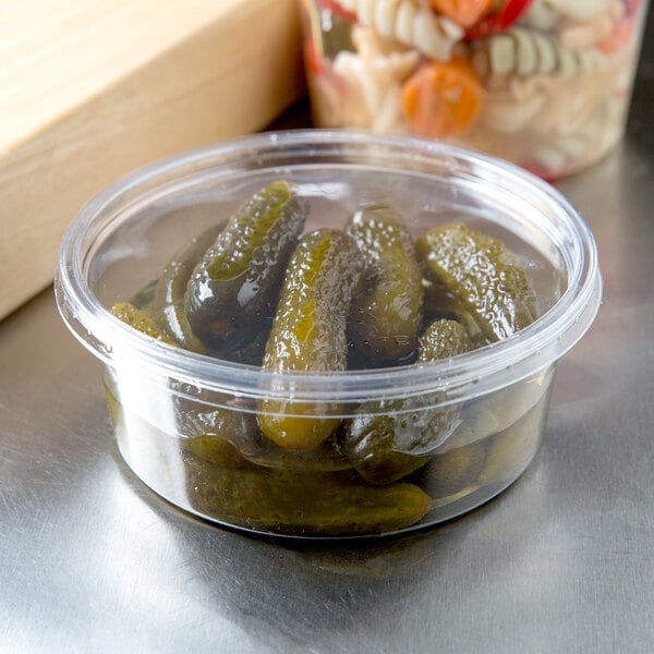 A plastic container of pickles next to a bowl of pasta on a counter in a deli.