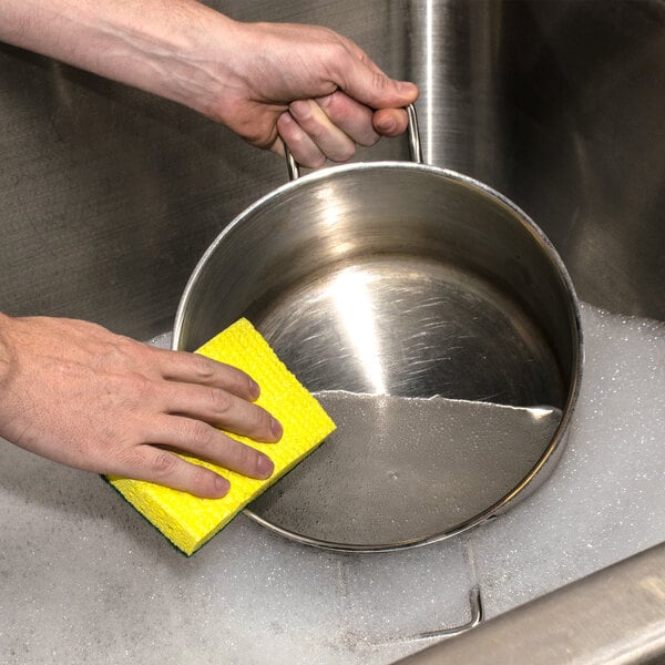 A hand holding a yellow 3M Scotch-Brite medium-duty scrub sponge washing a metal pan.