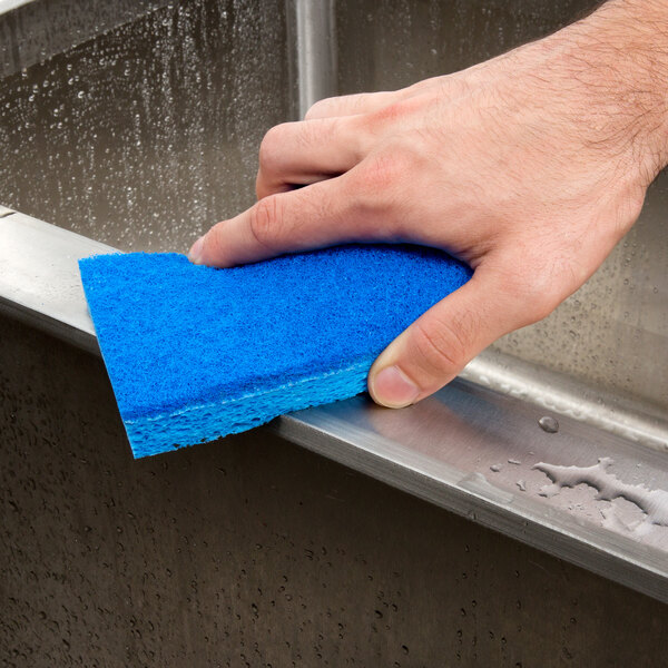 A hand using a blue 3M Scotch-Brite Soft Scour sponge to clean a metal sink.
