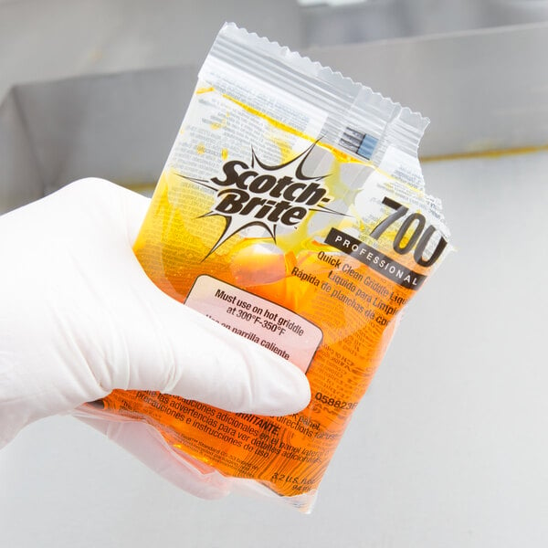 A gloved hand holding a 3M Scotch-Brite liquid griddle cleaning packet.