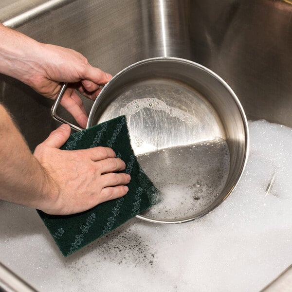 A hand using a 3M Scotch-Brite green scouring pad to wash a pot.