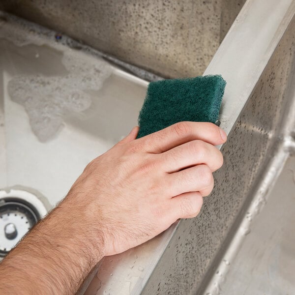 A hand using a 3M Scotch-Brite green scouring pad to clean a metal surface.
