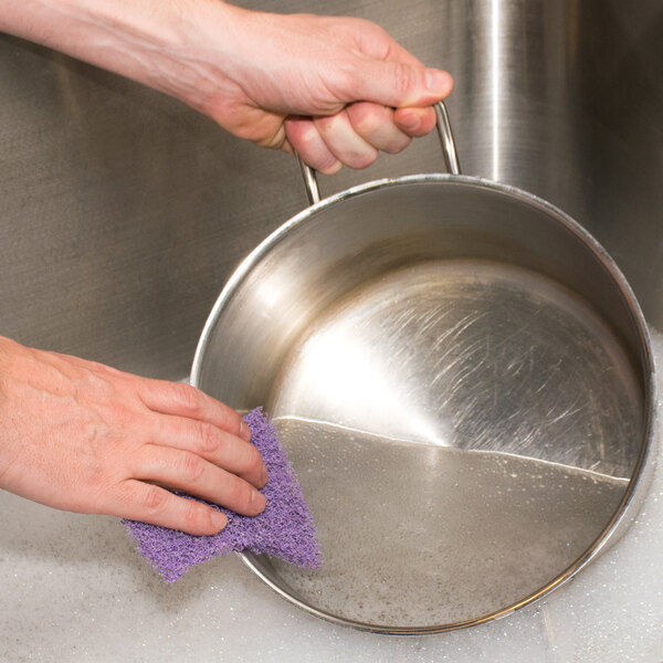 A person cleaning a stainless steel pan with a 3M Scotch-Brite purple scour sponge.
