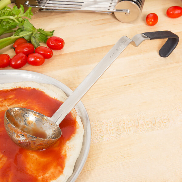 A Vollrath stainless steel ladle with a black handle on a plate with pizza dough and tomato sauce.