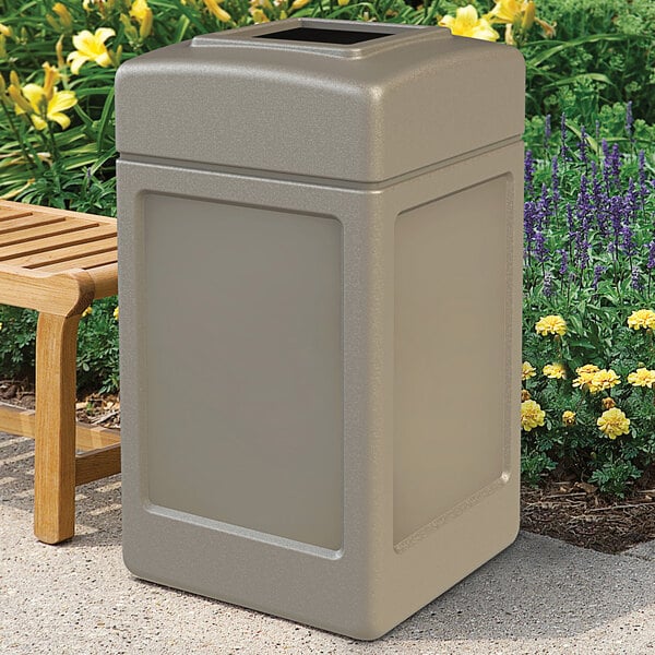 A beige Commercial Zone PolyTec waste container sitting on concrete next to a bench.