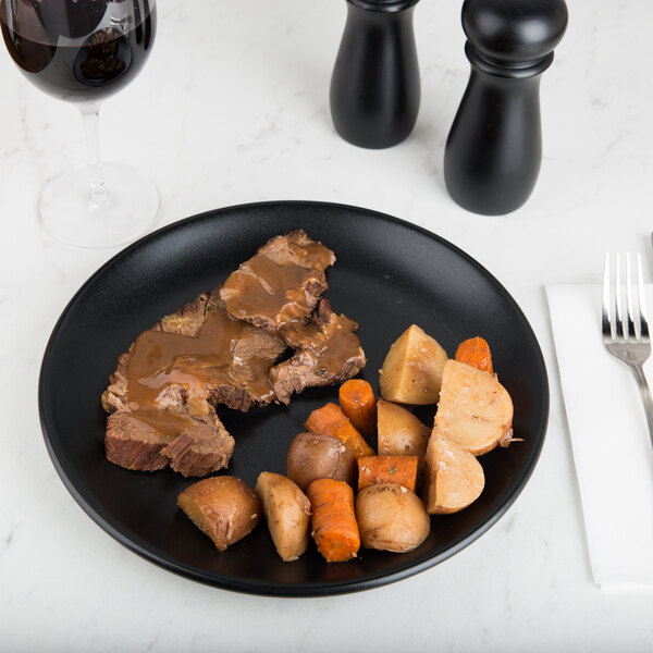 A Hall China Foundry black coupe plate with meat and vegetables on it next to a glass of red wine.