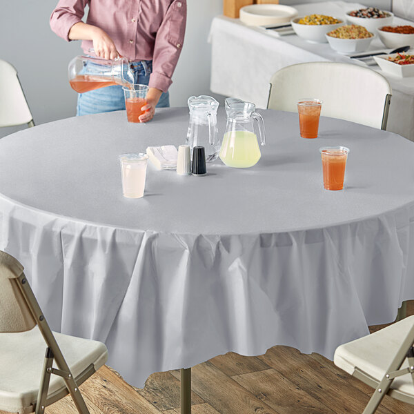 A woman pouring yellow liquid from a pitcher onto a white table with a Shimmering Silver OctyRound table cover and glasses of liquid.