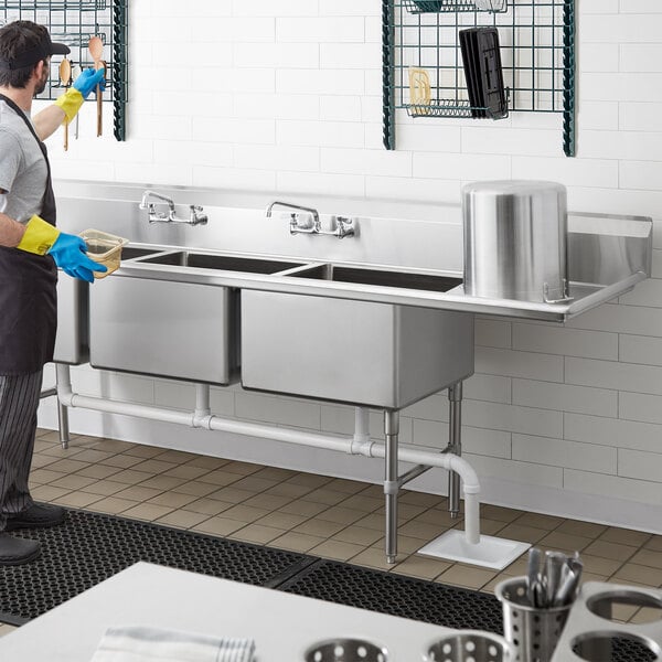 A man in an apron standing in front of a Regency stainless steel three compartment sink in a professional kitchen.