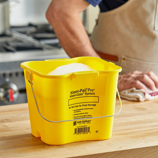 A man using a San Jamar yellow bucket with a wire handle to clean a professional kitchen counter.
