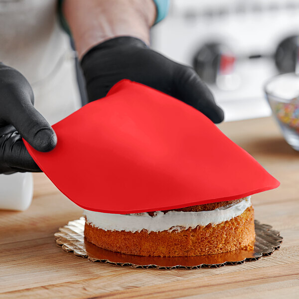 A person wearing black gloves and using a red spatula to spread Satin Ice red fondant over a cake.