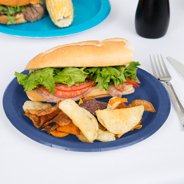 A sandwich with lettuce and tomato on a navy blue paper plate with potato chips.