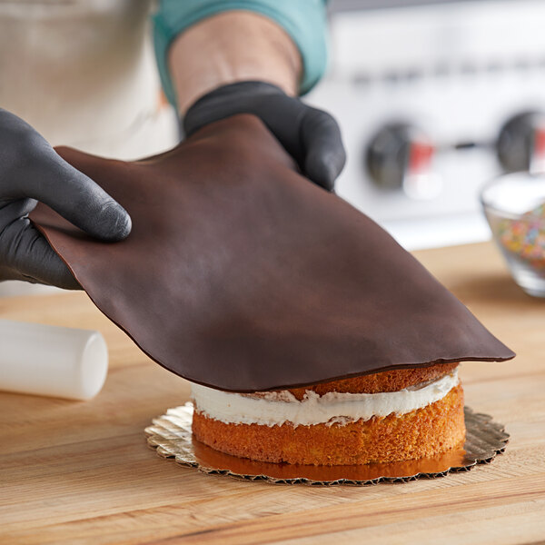 A person in gloves using Satin Ice Dark Chocolate Rolled Fondant to decorate a cake.