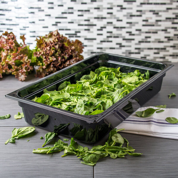 A Carlisle black plastic food pan filled with spinach on a counter.