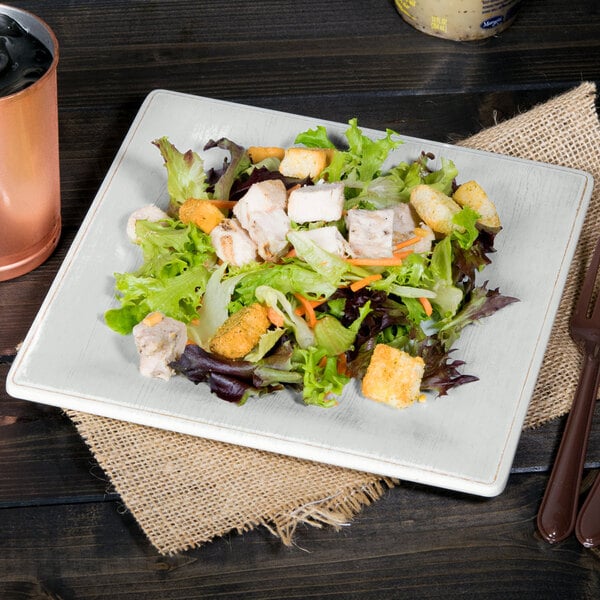 A white Carlisle melamine square salad plate with salad on it next to a copper cup.