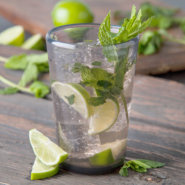 A Carlisle smoke Tritan plastic beverage glass filled with water, ice, and limes with a lime wedge on the table.