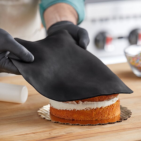 A person in black gloves using Satin Ice black rolled fondant to cover a cake.