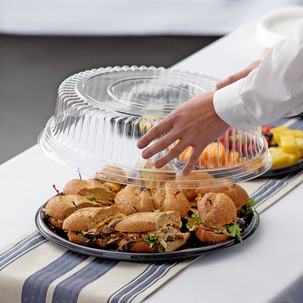 A person reaching for a sandwich on a clear plastic catering tray with a high dome lid.