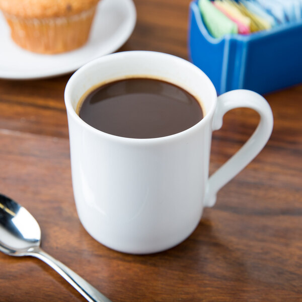 A white Arcoroc porcelain mug filled with brown liquid sitting on a table with a muffin.