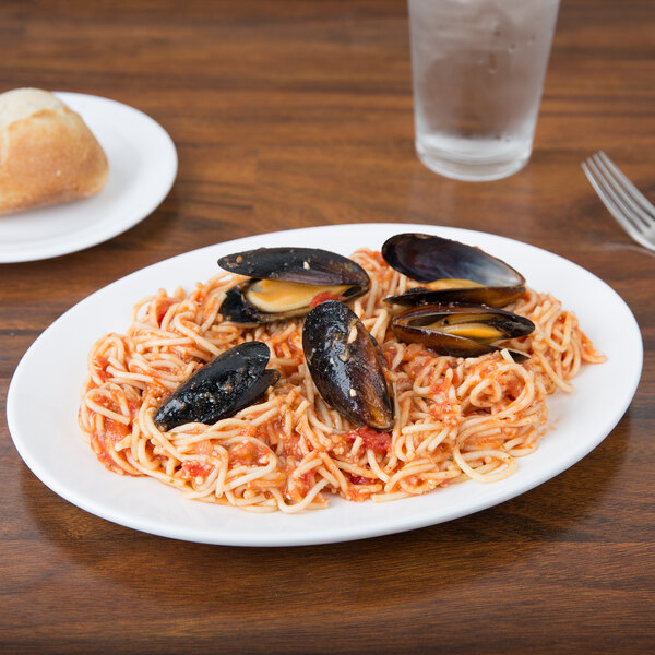 A Chef & Sommelier white bone china oval platter with spaghetti and mussels on it, with bread on the table.