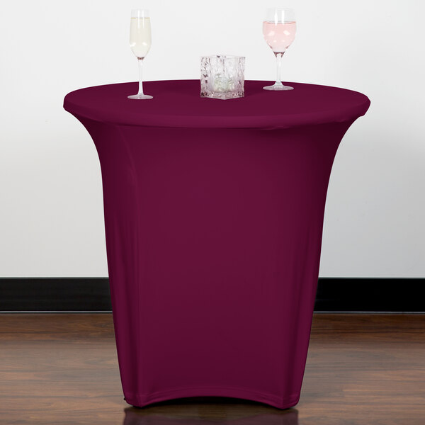 A table with a Snap Drape burgundy spandex round table cover and wine glasses on it.