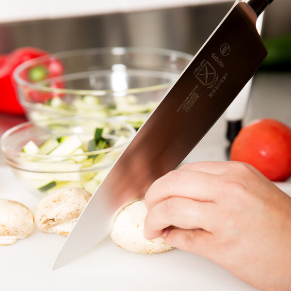 A person using a Mercer Genesis Forged Chef Knife to cut mushrooms on a cutting board.