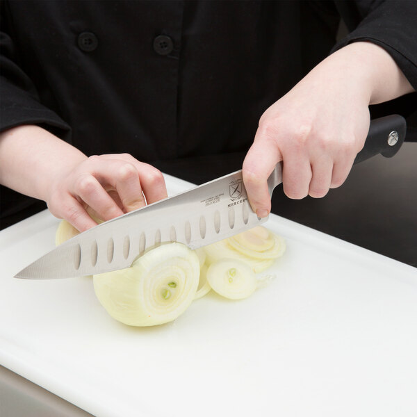 A person using a Mercer Culinary Genesis chef knife to slice onions on a white cutting board.