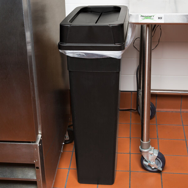A black rectangular Continental wall hugger trash can with a Drop Shot lid and plastic bag next to a stainless steel sink.