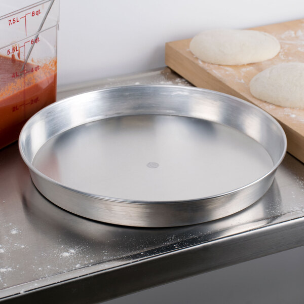 An American Metalcraft aluminum pizza pan on a table with dough and a wooden cutting board.