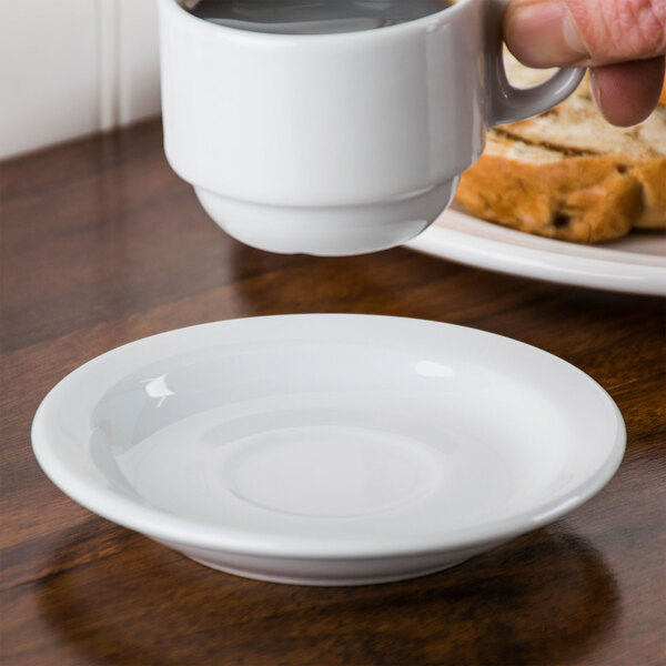 A hand holding a Tuxton demitasse cup of coffee over a white saucer on a table.