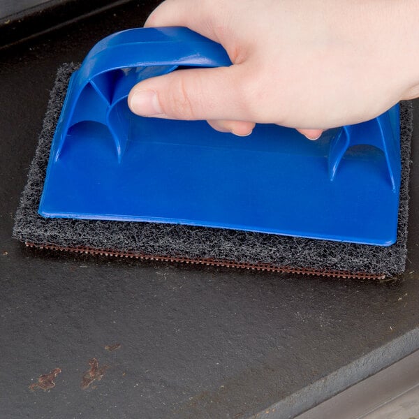 A hand using a blue-handled sponge to clean a grill.