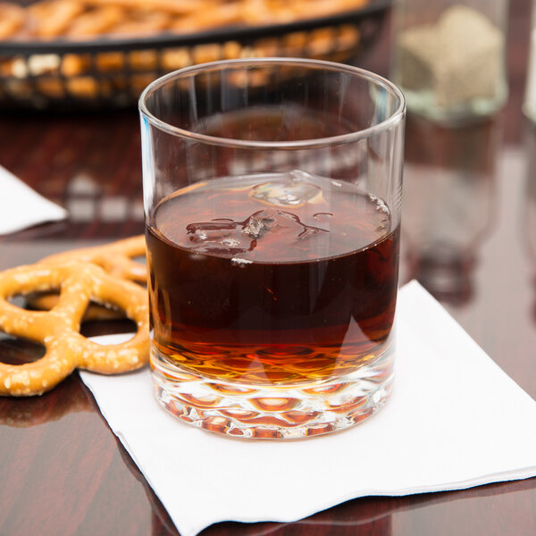 A Libbey customizable rocks glass filled with brown liquid on a table with pretzels.